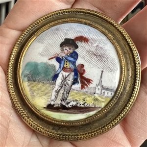 Painted porcelain button of a boy in blue playing a flute.