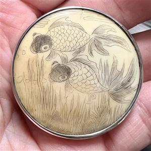 Incised, inked and painted natural material button of two fish swimming.