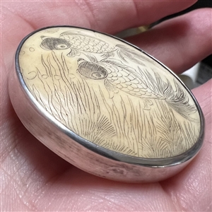 Incised, inked and painted natural material button of two fish swimming.